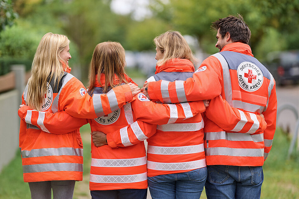 Vier Ehrenamtliche in Einsatzkleidung des Roten Kreuzes stehen mit dem Rücken zur Kamera und halten sich in den Armen.