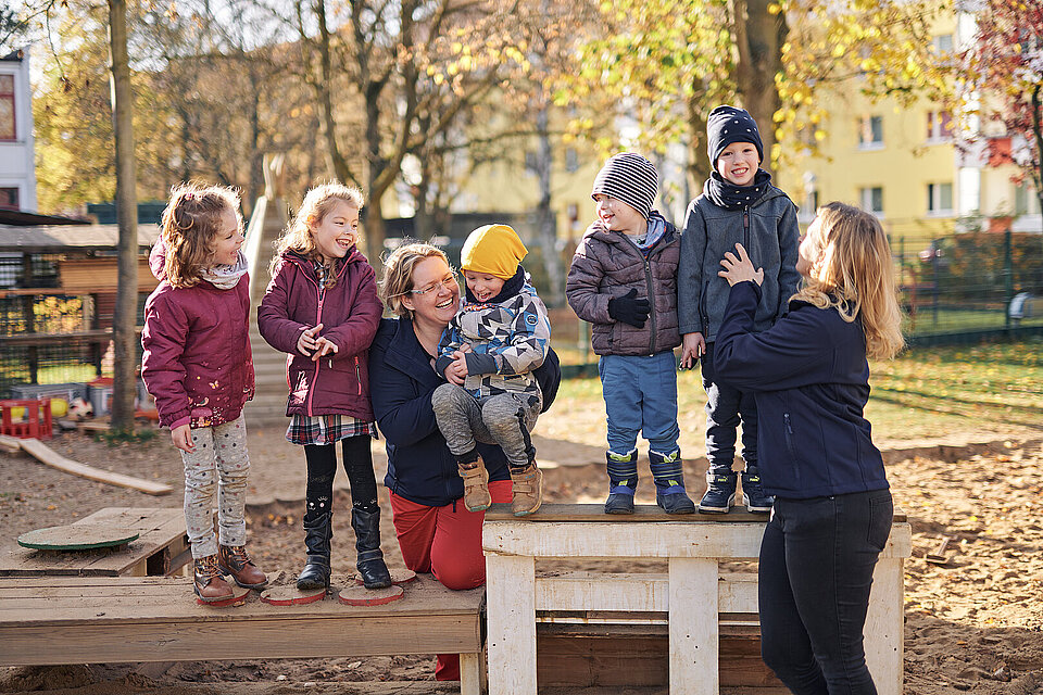 Zwei Erzieherinnen spielen mot fröhlich aussehenden Kindern auf einem Spielplatz.