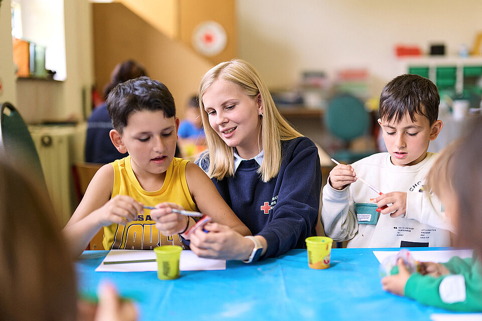 Eine junge Frau im Rotkreuz-Pulli malt mit zwei Kindern an einem Tisch sitzend.