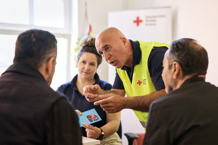 Ein Mann in einer Weste mit dem Logo des Roten Kreuz spricht gestikulierend mit zwei Männern, die mit dem Rücken zur Kamera sitzen. Im Hintergrund sitzt eine Frau, die ein Oberteil mit Rotkreuz-Logo trägt.
