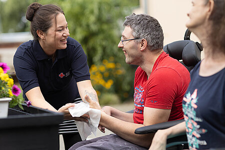 Eine Mitarbeiterin des Roten Kreuzes interagiert zugewandt mit einem Mann und eine Frau, die beide im Rollstuhl sitzen.
