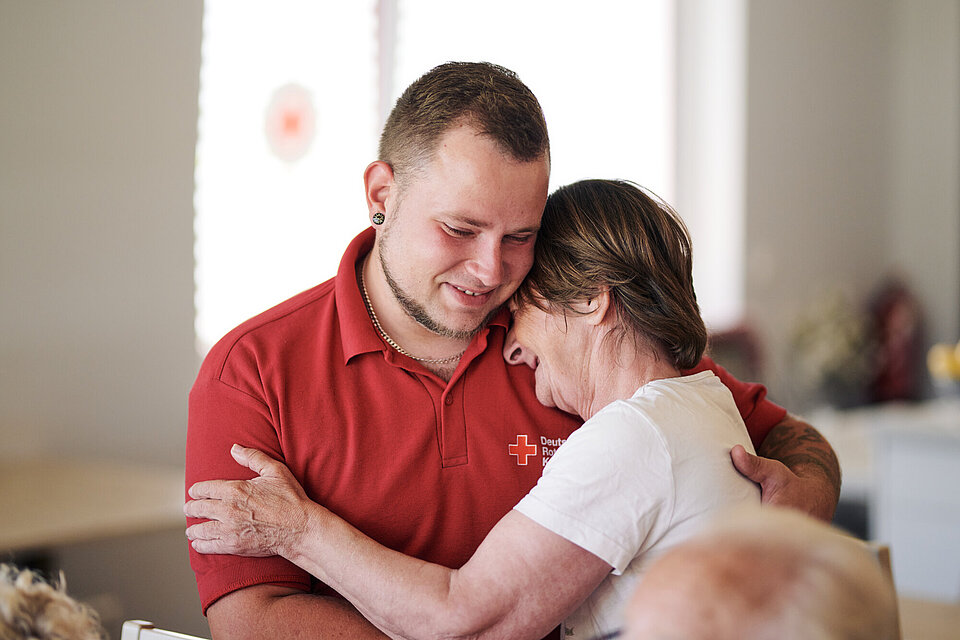 Ein Pfleger mit hält eine Seniorin im Arm. Der Pfleger trägt ein rotes T-Shirt des Roten Kreuzes.