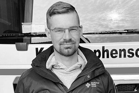 Porträtfoto in Schwarzweiß eines Ehrenamtlichen des Deutschen Roten Kreuzvor einem Einsatzwagen für den Katastrophenschutz steht und in die Kamera lächelt.