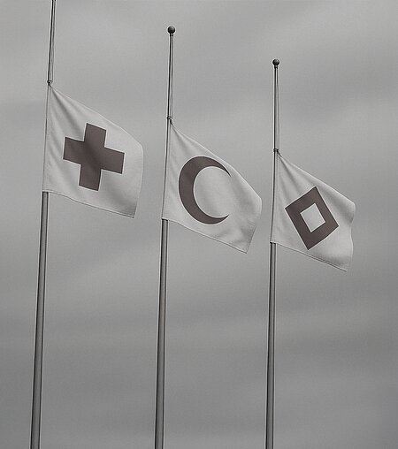 In schwarz-weiß hängen Flaggen des Roten Kreuzes, des Roten Halbmonds und des Roten Kristalls auf Halbmast (Bild: DRK).