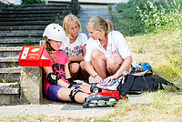 Frau verarztet ein Mädchen nach einem Rollerscateunfall. Ein zweites Kind sieht zu.