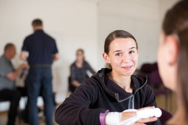 Junge Frau schaut in die Kamera. Ihr wird gerade ein Verband an der Hand angelegt.