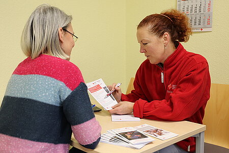 Eine Frau in einer Jacke des Rotkreuz-Suchdienstes zeigt einer anderen Frau eine Informationsbroschüre.