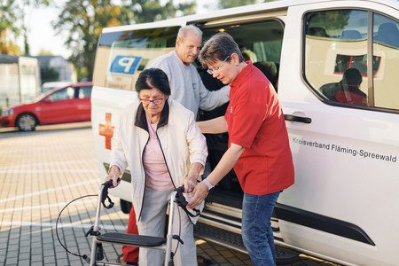 Eine Frau und ein Mann helfen einer Seniorin mit Rollatur aus einem Transporter des Fahrdienstes des Roten Kreuzes.