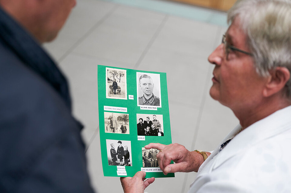 Eine ältere Frau zeigt einem Mann schwarz-weiß Bilder, die Menschen abbilden..