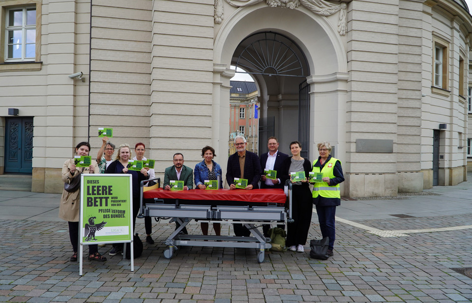 Vertreter*innen der Spitzenverbände der Freien Wohlfahrtspflege in Brandenburg stehen vor dem Landtag um ein symbolisches leeres Pflegebett.
