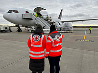 Zwei Menschen in DRK-Kleidung stehen auf einem Flugfeld und beaobachten die Beladung eines Flugzeugs