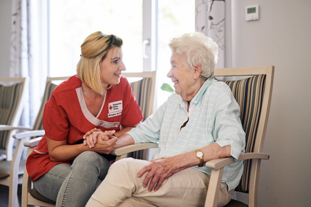 Eine junge Pflegerin in DRK-Kleidung sitzt zugewandt neben einer Seniorin und hält deren Hand. Beide lächeln.