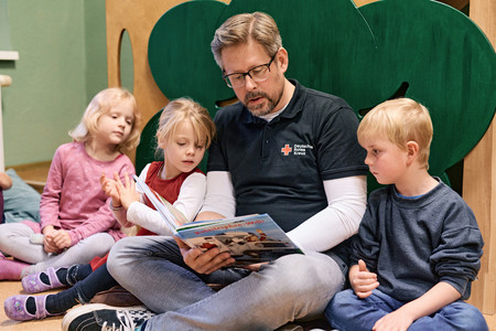 Ein Erzieher mit einem Rotkreuz-Logo auf dem T-Shirt sitzt in der Mitte von drei Kindern und liest ihnen aus einem Buch vor.