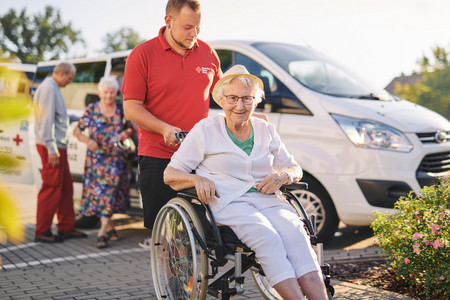 Ein Mitarbeiter des Roten Kreuz schiebt eine fröhliche Frau im Rollstuhl. Im Hintergrund ist ein Fahrzeug des Roten Kreuz zu sehen.