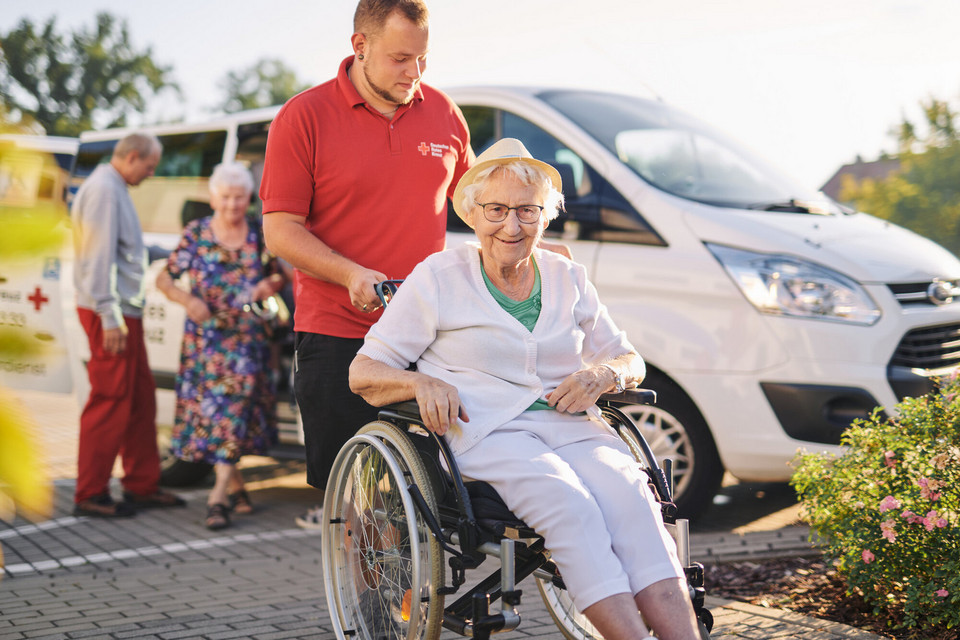 Ein Mitarbeiter des Roten Kreuz schiebt eine fröhliche Frau im Rollstuhl. Im Hintergrund ist ein Fahrzeug des Roten Kreuz zu sehen.