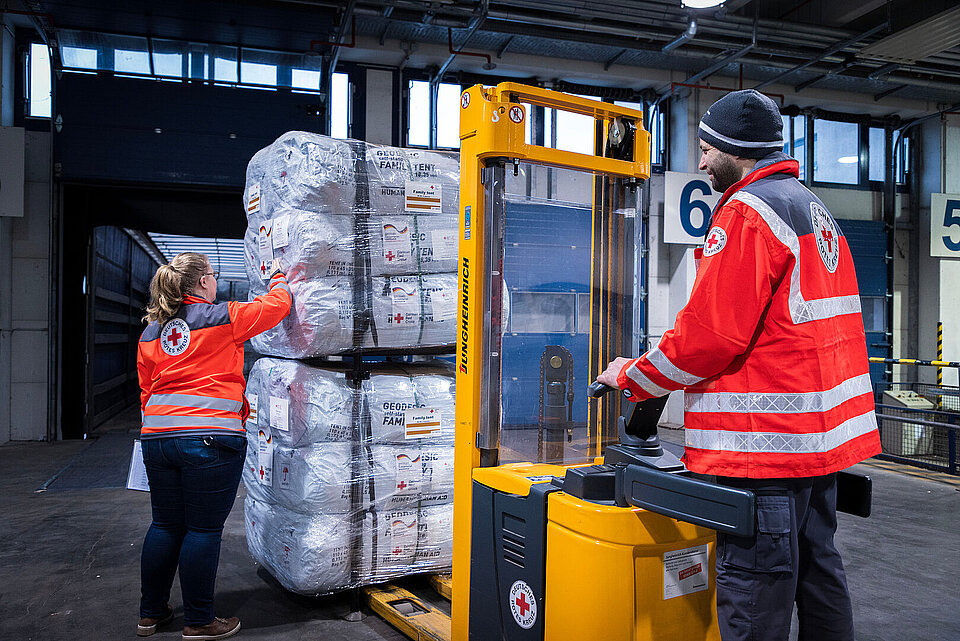 Eine Frau und ein Mann in Einsatzkleidung des Roten Kreuzes verladen Hilfsgüter mit einem Gabelstapler.