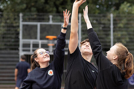 Drei junge Menschen halten jeweils einen Arm in die Höhe und deuten ein Basketballspiel an.