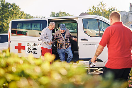Mitarbeitende des DRK helfen einem älteren Herrn aus einem DRK-Fahrzeug.